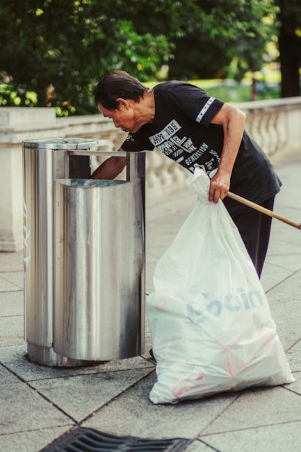 An overflowing collection of mixed household waste and recycling, including black and black and blue bins, cardboard boxes, paper, plastics, and various discarded items, situated on a paved urban stre