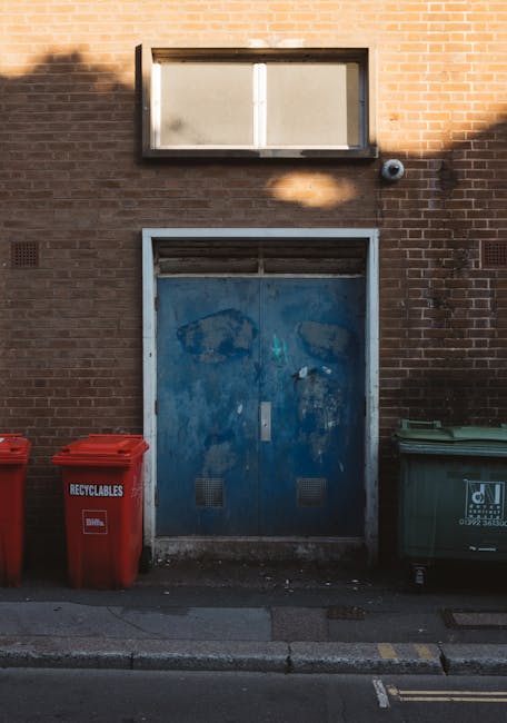 A collection of overflowing rubbish bins and scattered waste on a paved urban sidewalk, with a large grey mixed paper and cardboard bin positioned centrally, partially filled with crumpled papers, car